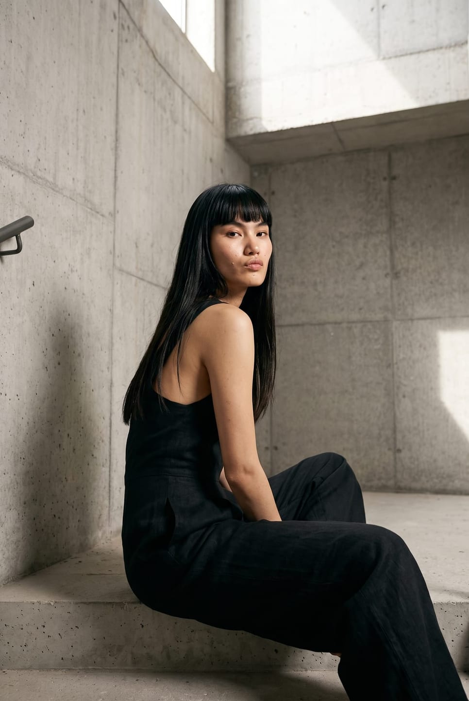 Synthetic talent in a black jumpsuit, seated against a raw concrete wall — not a real person